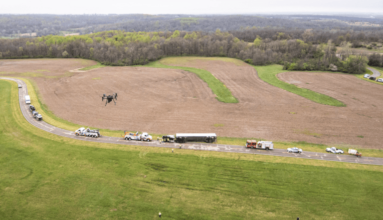 A black drone hovers above a tractor trailer accident on a windy two-lane road with grassy fields on either side.