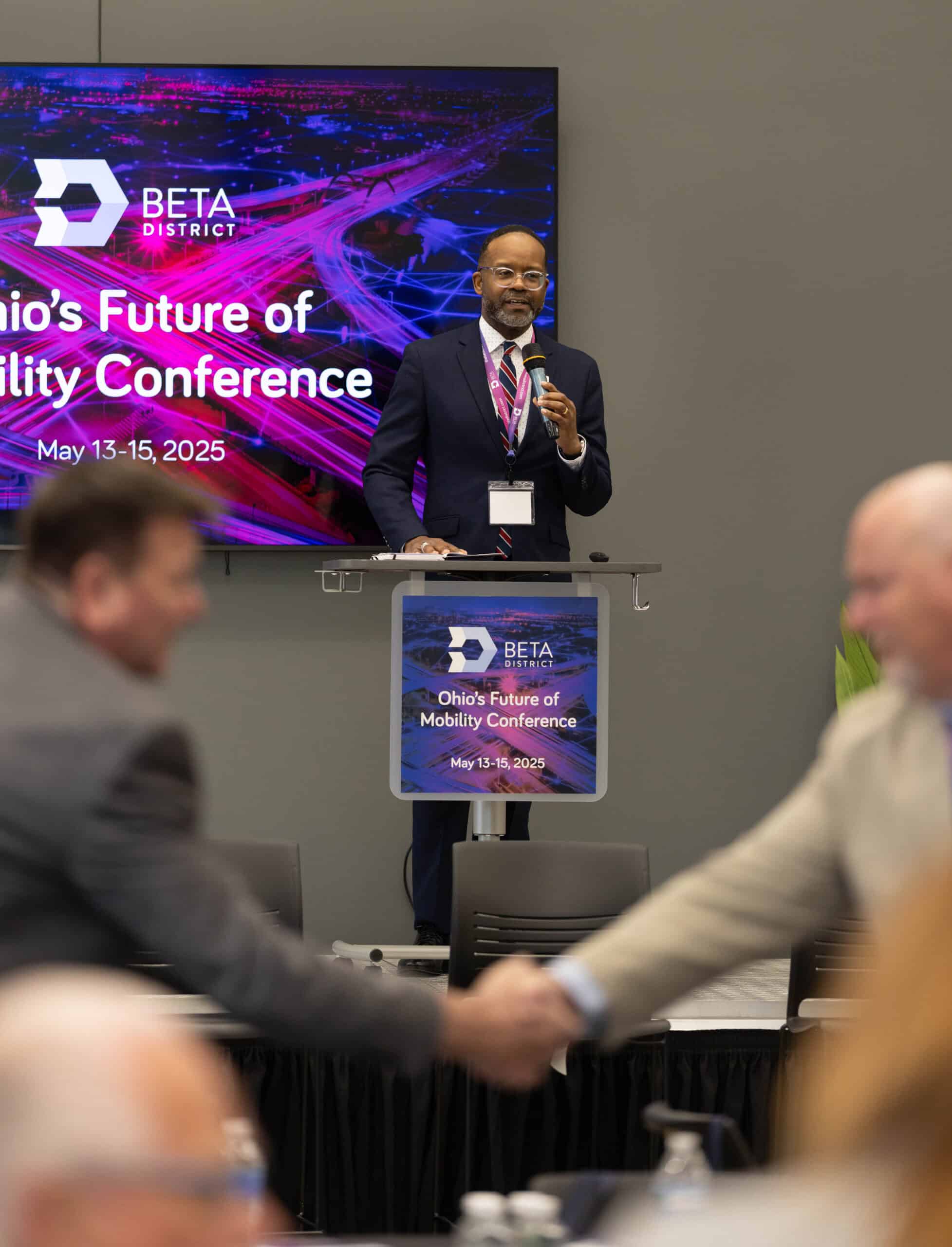 Two individuals shaking hands and networking at the inaugural Ohio's Future of Mobility conference. This event took place in The Beta District. Executive Director Doug McCollough is on the stage speaking to kick off the event.