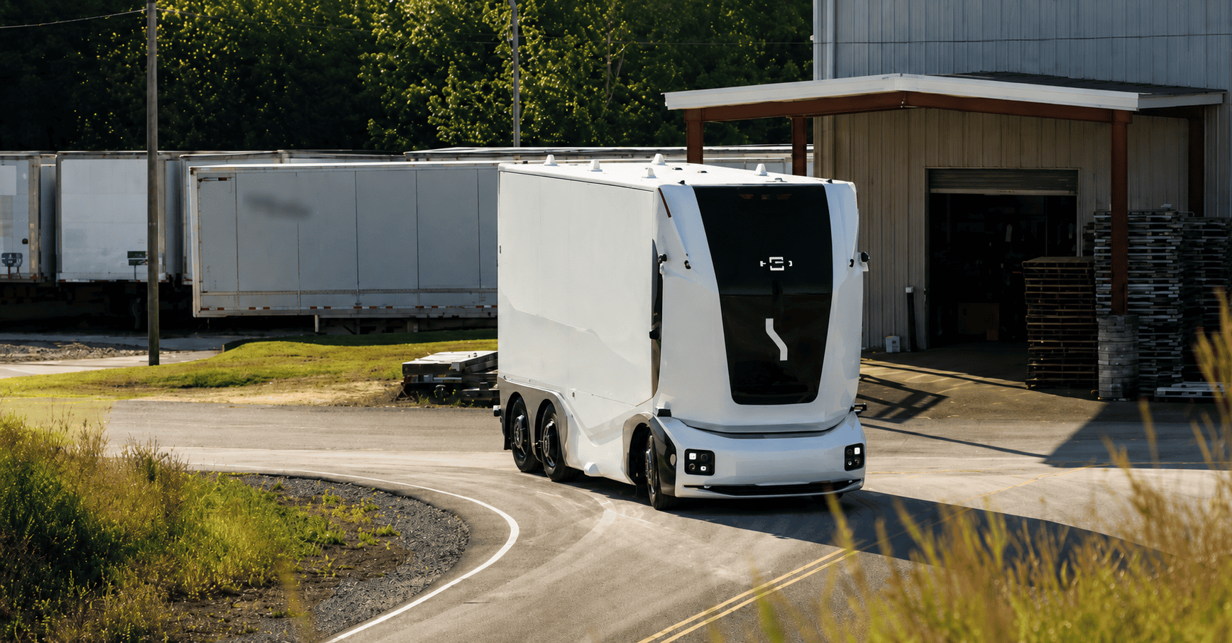 A white autonomous electric truck drives along a curved road outside a warehouse loading area, with trailers and greenery in the background.